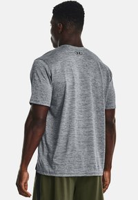 Muž oblečený v šedém tričku s krátkým rukávem sportovního typu s malým logem na horní části zad a olivově zelených šortkách, který je otočen zády k fotoaparátu.