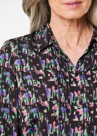 Camicia nera a bottoni con un motivo astratto colorato in verde, viola e rosa; presenta un colletto e pieghe verticali lungo la parte anteriore.