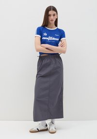 Blauw kortemouwen shirt met witte accenten en tekst, gecombineerd met een lange grijze nylon rok. Het model draagt witte sneakers met marineblauwe details.