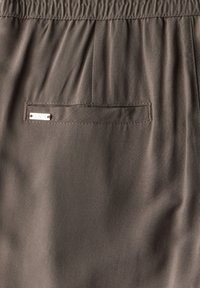 Brown fabric with a smooth texture features a rear pocket and elastic waistband. A small silver tag is stitched onto the pocket.