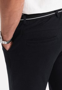 Zwarte textuur broek met een slim fit, zijzakken en een contrasterende witte streep op de tailleband. Subtiele details op de achterzak.