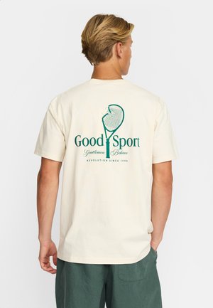 Beige bomulds t-shirt med et grønt grafisk print af en tennisketsjer og tekst på bagsiden. Kortærmede og med rund halsudskæring.