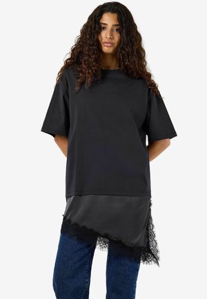 Kvinde med krøllet hår iført en løs sort t-shirt over et sort slip-nederdel med blonder og blå jeans, står med hænderne bag ryggen.