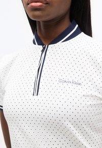 Κοντινό πλάνο ενός ατόμου που φοράει λευκό πόλο πουκάμισο με μικρές μαύρες βούλες, ναυτικό γιακά και φερμουάρ, καθώς και λογότυπο "Calvin Klein" στο στήθος.