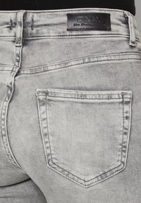 Jeans de mezclilla gris que presentan una vista cercana de la parte trasera con un bolsillo visible, detalles de costura sutiles y un parche de cuero negro con el logo.