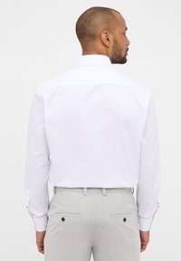 Camicia bianca a maniche lunghe con colletto standard, dotata di polsini con bottoni, abbinata a pantaloni leggeri grigi a motivo, mostrata da dietro.
