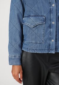 Quiltet denimjakke i lys blå farge, med trykknapper og en brystlomme. Ermet har en brettet mansjettdetalj.