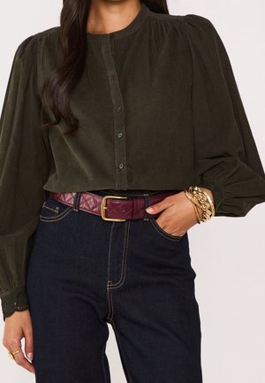 Frau trägt dunkelgrüne, geknöpfte Bluse mit Puffärmeln, dunkle Jeans, bordeauxfarbenen Gürtel mit goldener Schnalle und goldene Armbänder am rechten Handgelenk.