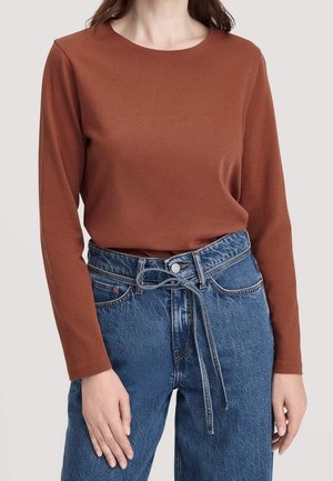 Vrouw draagt een roestkleurige top met lange mouwen, ingestopt in een lichtblauwe, hooggetailleerde jeans met een gebonden spijkergordel.