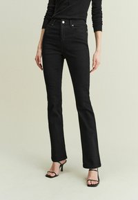 Jeans neri a zampa d'elefante realizzati in denim resistente, con vita alta, chiusura a bottone e dettagli di cucitura discreti. Abbinati a tacchi alti neri con cinturini.