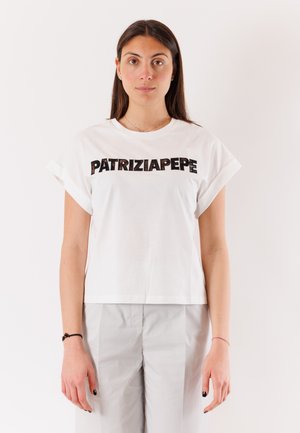 Donna con lunghi capelli scuri che indossa una maglietta bianca con la scritta "PATRIZIA PEPE" e pantaloni grigio chiaro, in piedi davanti a uno sfondo uniforme.