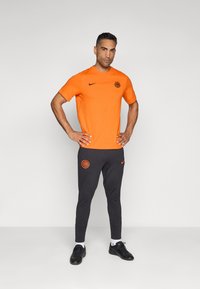 Oranža Nike īsām piedurknēm krekls ar melnu logotipu, apvienots ar pelēkām šaurām sporta biksēm ar oranžu logotipu un melnām sporta kurpēm.