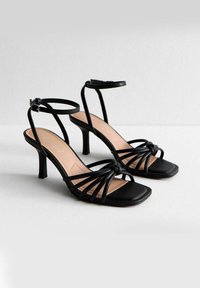 Czarne sandały na wysokim obcasie z kwadratowym noskiem, z cienkimi paskami i węzłem z przodu. Gładka faktura, minimalistyczny design.