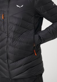 Giacca nera isolata con design trapuntato, dotata di zip frontale e tirazip arancioni. Logo bianco sulla spalla e vestibilità slim.