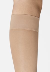 Beige strumpor som når låret med en slät yta och ett brett elastiskt band upptill. Genomskinligt material sträcker sig till mitten av låret.