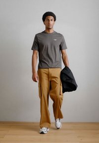 Pelēka īsām piedurknēm T-kreklu, brūnās krāsas kargo bikses ar sānu kabatām un melnu jaku, valkājot baltas un pelēkas sporta apavus, stāv uz koka grīdas.