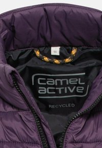 Fioletowa pikowana kurtka z czarną podszewką, logo "camel active", metką z rozmiarem 50 oraz oznaczeniem materiału z recyclingu wewnątrz kołnierza.