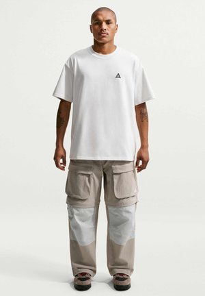 Man staat met het gezicht naar voren, draagt een wit T-shirt met een klein zwart logo en beige cargobroek met grote voorzakken en kniestukken.