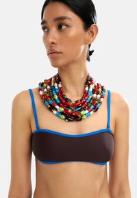 Wielowarstwowy naszyjnik z koralików, zawierający kolorowe okrągłe i cylindryczne koraliki w kolorach czerwonym, niebieskim, zielonym, brązowym i białym, umieszczony na neutralnym tle.