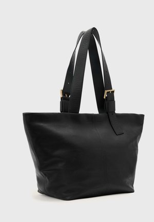 Zwarte leren shopper met twee verstelbare schouderbanden met gespen in goudkleur, tegen een effen lichtgrijze achtergrond.