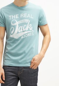 Světle modré tričko z bavlny s krátkými rukávy, na kterém je bílý potisk s logem "THE REAL Originals Jack & Jones." Klasický střih.