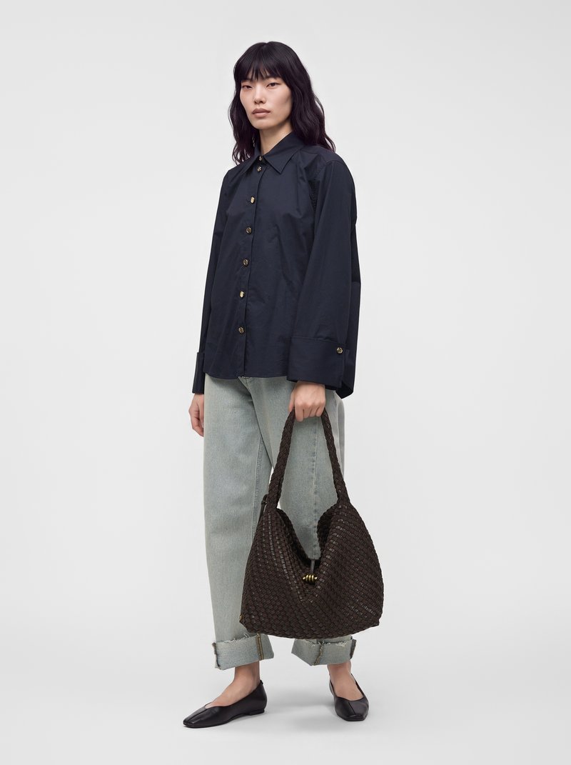 Femme aux longs cheveux noirs porte une chemise noire boutonnée et un jean clair, tenant un grand sac à main tissé foncé avec des poignées tressées.
