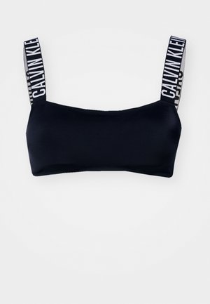 Črn modrček z gladko teksturo, opremljen s širokimi naramnicami, okrašenimi z belim napisom "CALVIN KLEIN". Minimalističen dizajn, pravokotna oblika.