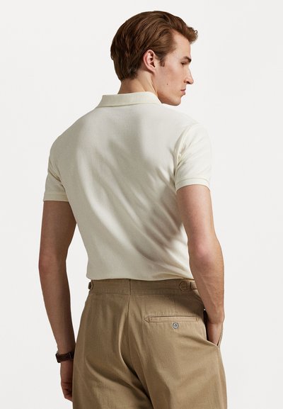 Kortermet kremfarget polo skjorte med flat krage, sammen med brune bukser med baklomme og beltehemper. Myk tekstur, tettsittende design.