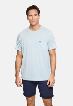 Hombre de pie mirando al frente, vestido con una camiseta de manga corta azul claro con bolsillo en el pecho y pantalones cortos azul marino remangados.