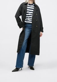 Černý dlouhý trenčkot se zlatými knoflíky, nošený přes námořnickou a bílou pruhovanou ko šili a široké džíny, doplněný černými jehlovými podpatky.