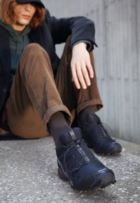 Young person with long hair wearing black cap, green hoodie, brown pants, and black trail running shoes sitting outdoors on concrete steps.
