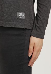 Langärmliges, dunkelgraues T-Shirt aus strukturiertem Stoff mit einem Logopatch am Saum. Das Shirt wird mit schwarzen Hosen kombiniert.