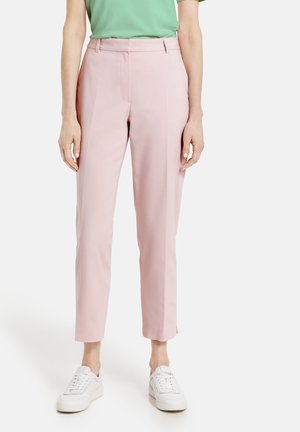 Persoon draagt lichtroze op maat gemaakte enkellange broek met zijsplits, gecombineerd met witte sneakers en een groene top.
