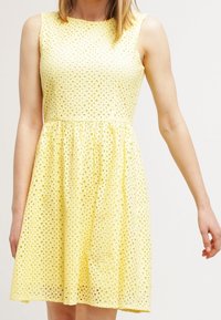 Vestido amarillo sin mangas con un patrón de ojal texturizado, corpiño ajustado y falda acampanada, que muestra una tela ligera y transpirable.