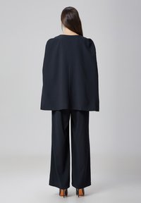 Haut de style cape noir avec des manches larges, associé à un pantalon noir taille haute et des chaussures à talons. Tissu lisse, design minimaliste.