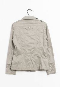 Giacca beige aderente in materiale di cotone leggermente testurizzato. Presenta spalline sulle spalle e polsini abbottonati. La parte posteriore ha una cucitura centrale con un bottone.