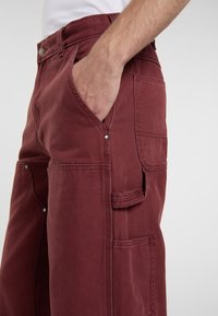 Pantaloni da lavoro bordeaux realizzati in tessuto resistente; dotati di tasche laterali, dettagli in metallo argentato e una vestibilità comoda con cuciture a vista.