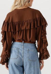 Vrouw in een bruine ruffled blouse met lange mouwen, ingestopt in lichtblauwe denim jeans met hoge taille, achterzakken en een leren riem.