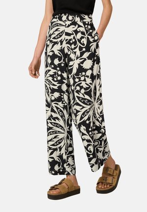 Femme portant un pantalon large à fleurs noir et blanc avec poches, associé à des sandales marron à deux brides à plateforme et un top noir sans manches.