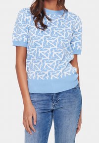 Světle modrý svetr s krátkými rukávy, který má bílý abstraktní vzor. Hladký žebrovaný výstřih a lem, vyrobený z měkké, strukturované tkaniny.