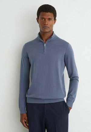 Mand iført en lyseblå langærmet lynlås-sweater og mørke bukser står op ad en ensfarvet hvid væg.