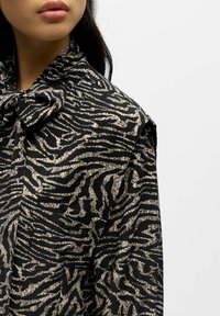 Musta pusero, jossa on beige teksturoitu seeprajuoviointi. Siinä on rusetti kauluksessa ja laskeutuva kangas, joka näyttää hieman kiiltävältä.