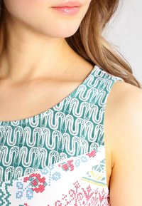 Monivärinen mekko, jossa on vihreä kuviollinen yläosa valkoisilla aalto kuvioilla, siirtyen valkoiseen alaosaan kukkakuosisilla. 