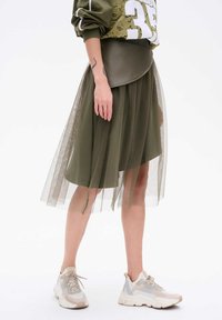 Spódnica z warstwowego tiulu w oliwkowym kolorze, z skórzanym pasem, połączona z jasnymi sneakersami. Materiał charakteryzuje się przezroczystą teksturą i zwiewnym krojem.