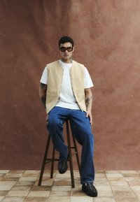 Egy fiatal férfi tetoválásokkal, napszemüveget visel, bézs mellényt, fehér pólót, kék farmert és fekete cipőt visel, egy fa széken ül barna fal előtt.