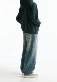 Personne portant un sweat à capuche vert foncé, un jean bleu délavé ample et des baskets blanches, debout devant un fond blanc uni.