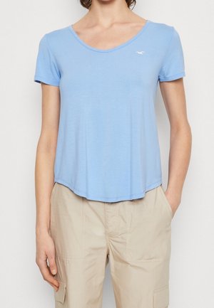 Kvinde iført en lyseblå T-shirt med korte ærmer og V-hals samt beige cargobukser med hænderne afslappet langs siderne mod en ensfarvet baggrund.