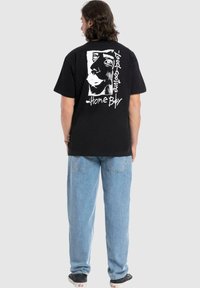 Zwarte katoenen T-shirt met een witte grafiek van een gezicht en tekst op de achterkant. Model gecombineerd met lichtblauwe denim broek en zwarte sneakers.
