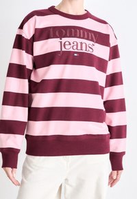 Sudadera a rayas granate y rosa con cuello redondo, que lleva un bordado con las palabras "tommy jeans" y un pequeño detalle de logo en la parte delantera.