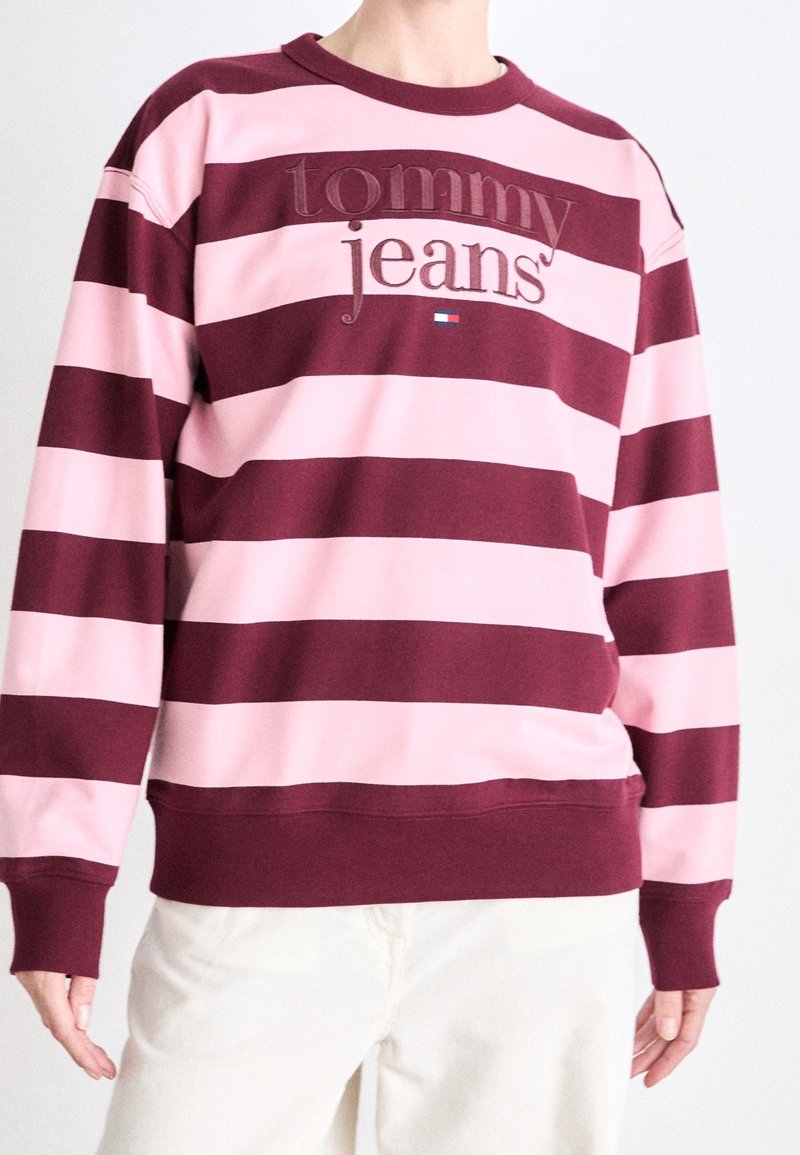 Sudadera a rayas granate y rosa con cuello redondo, que lleva un bordado con las palabras "tommy jeans" y un pequeño detalle de logo en la parte delantera.